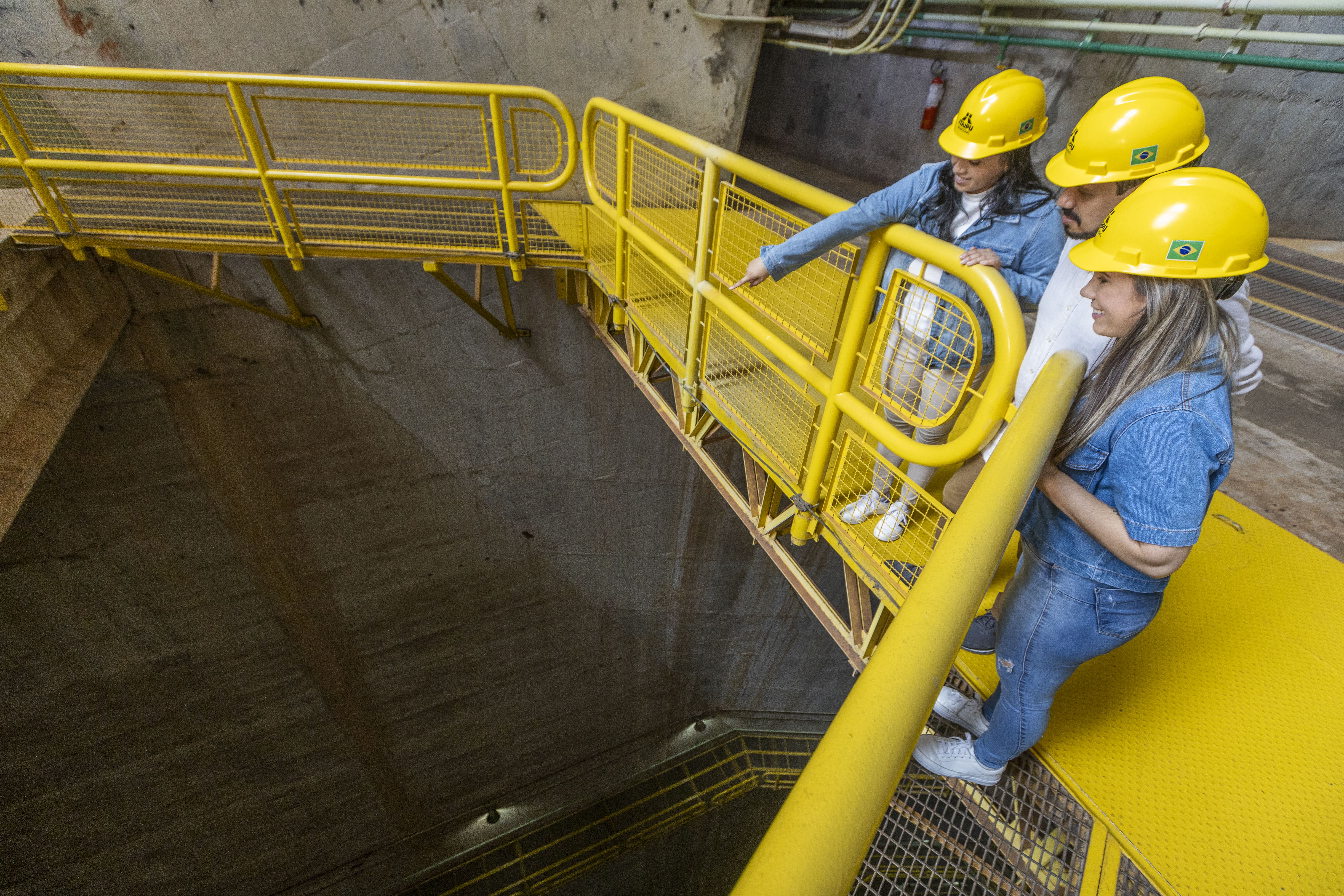 Três pessoas usando capacete amarelo, duas mulheres e um homem, dentro da Usina Hidrelétrica de Itaipu, localizada na em ambos os países Brasil e Paraguai.