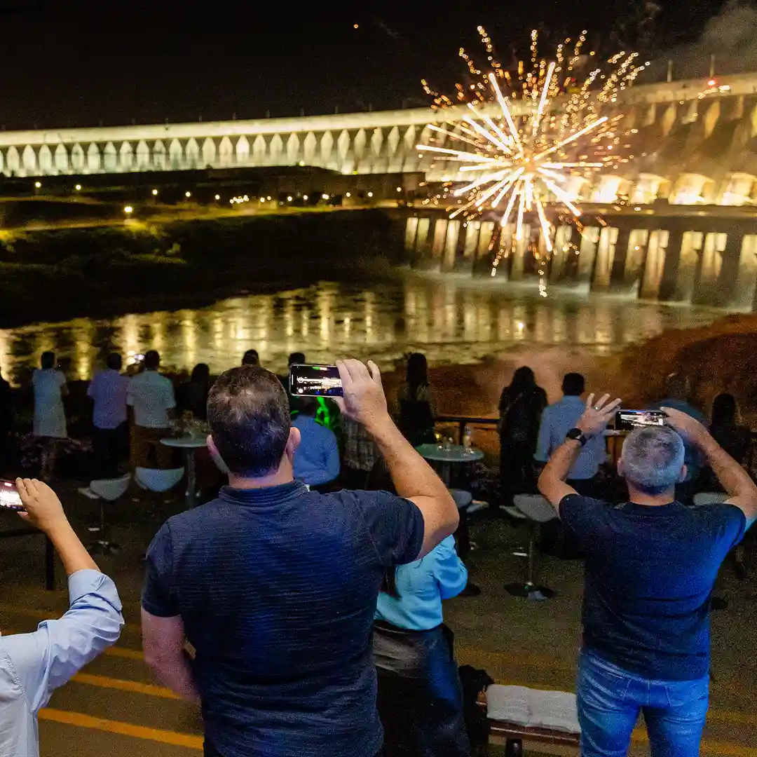 Pessoas tirando foto da iluminação da barragem em evento no Mirante Central na Usina Hidrelétrica de Itaipu, com fogos de artifício no fundo.