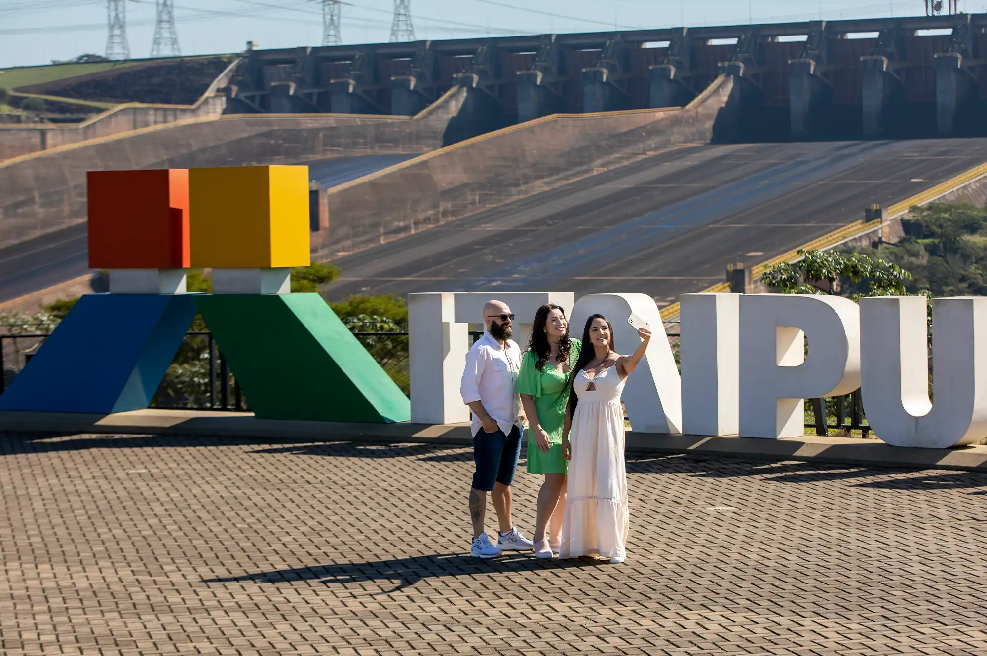 Duas mulheres, uma tirando a foto, e um homem, posando em frente ao letreiro da Usina Hidrelétrica de Itaipu, localizada em Foz do Iguaçu, na divisa entre o Brasil e Paraguai.