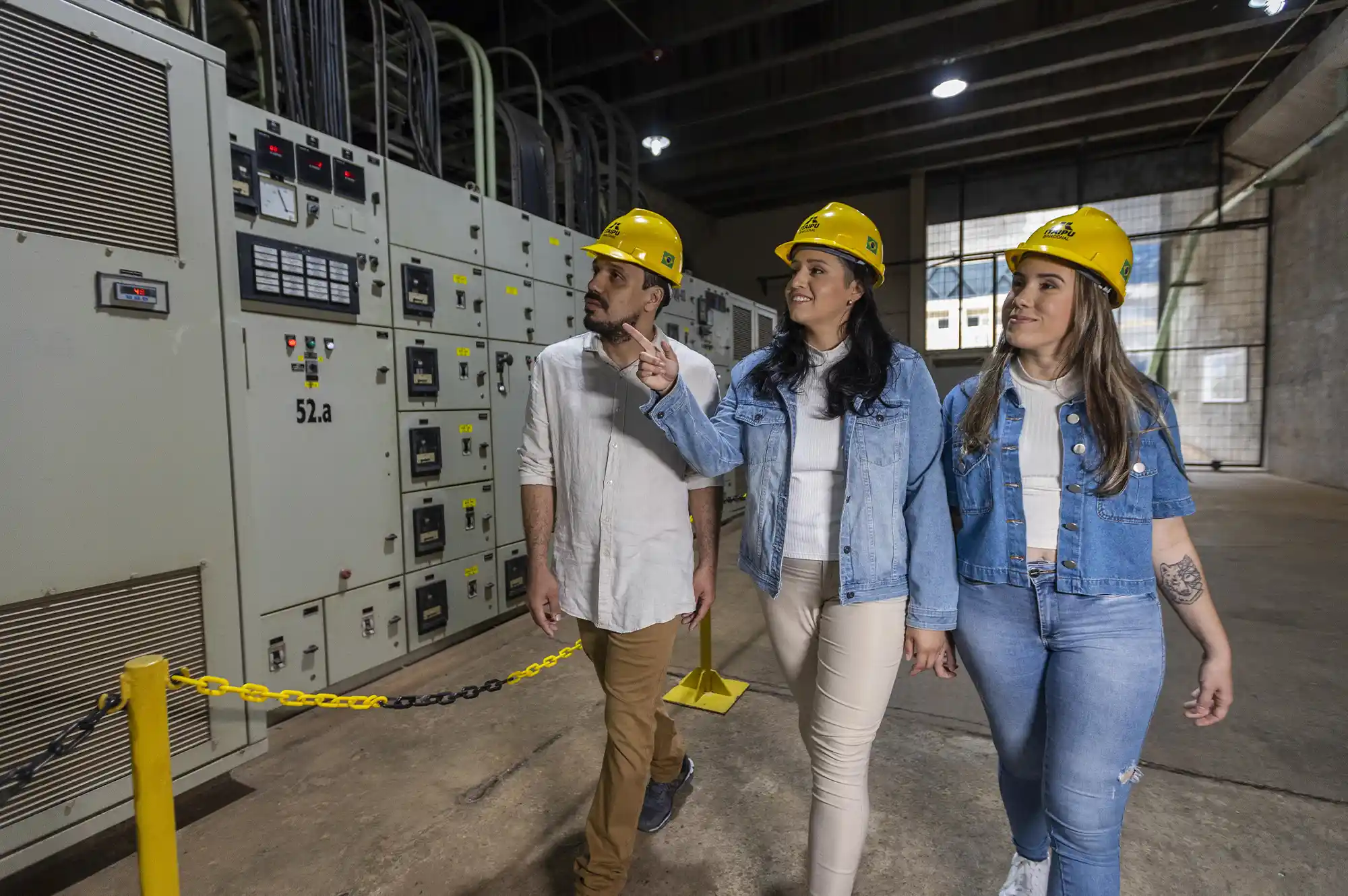 Um homem e duas mulheres usando capacete amarelo dentro da Usina Hidrelétrica de Itaipu, passando por máquinas.