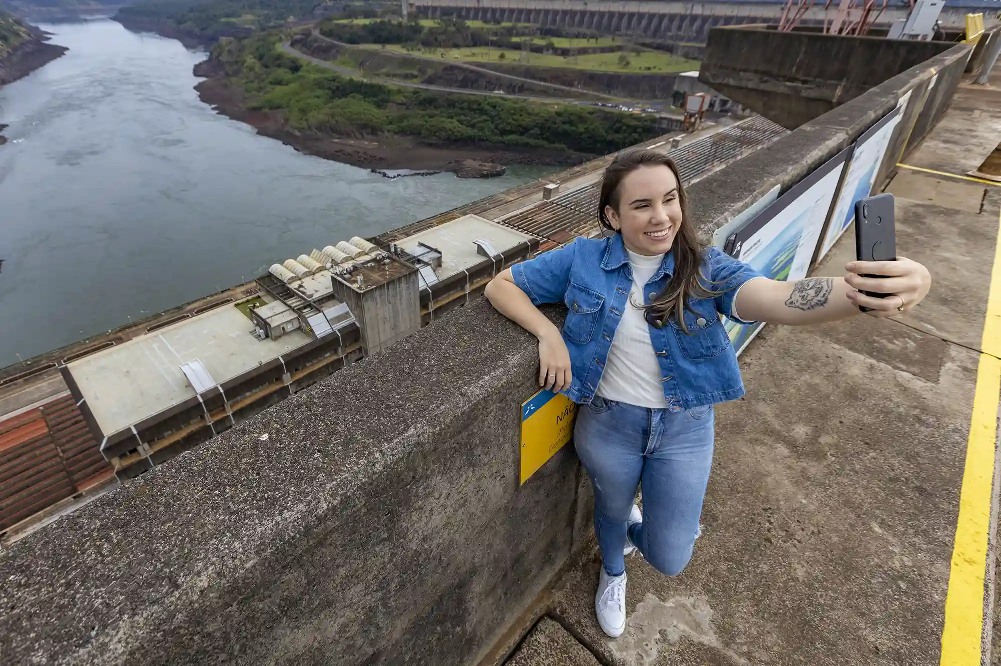 Mulher tirando selfie no topo da Barragem da Usina Hidrelétrica de Itaipu, localizada em Foz do Iguaçu, com vista para o Rio Paraná e o lado do Paraguai da usina.