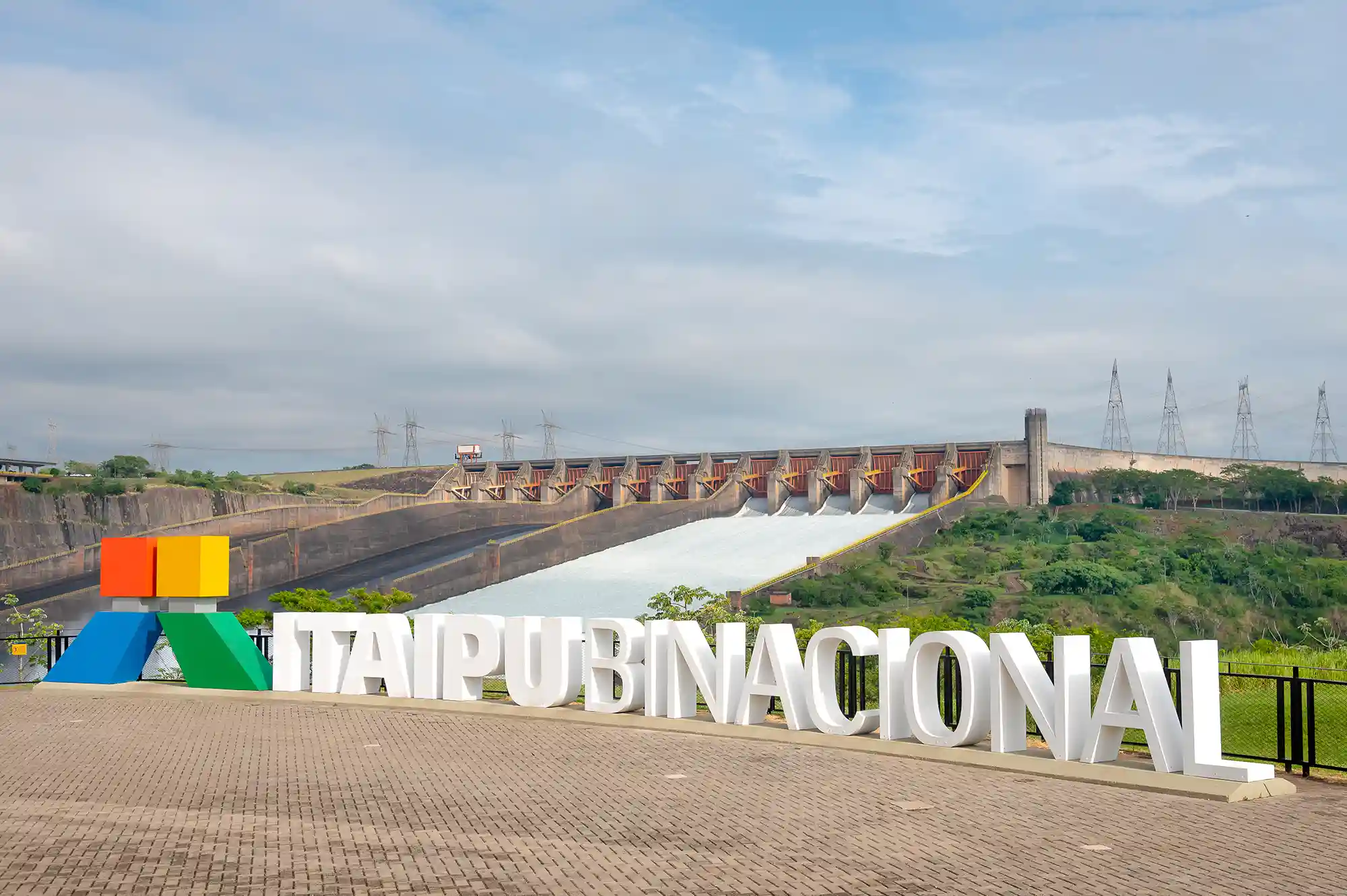 Letreiro da logo da Usina Hidrelétrica de Itaipu, localizada em Foz do Iguaçu, com o vertedouro aberto ao fundo e céu nubaldo.